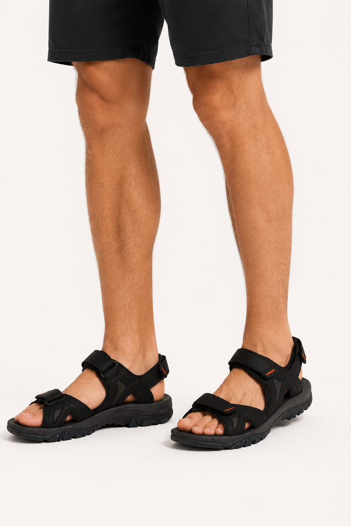 Man draagt zwarte sportieve sandalen en korte broek, staand op een witte achtergrond.