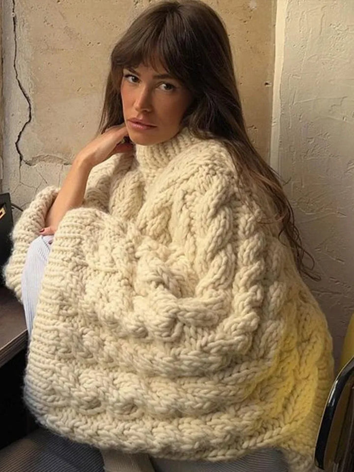 Vrouw in oversized witte gebreide trui met kabelpatroon zit in een fauteuil, comfortabel en stijlvol.