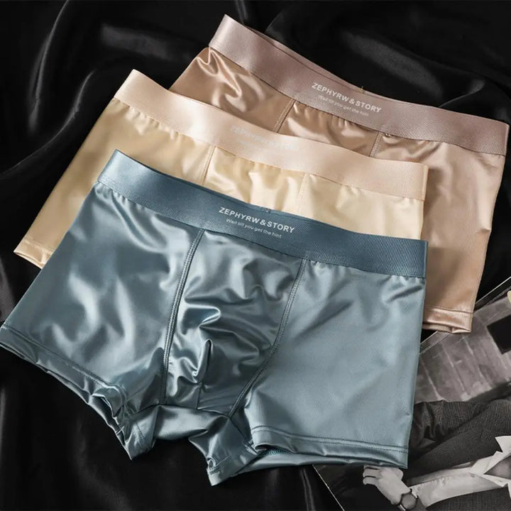 Drie glanzende herenboxershorts in blauw, beige en bruin op een zwarte achtergrond.