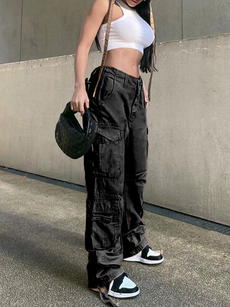 Vrouw in een zwarte cargobroek met wijde pijpen, witte crop top en zwarte tas, poseert casual.