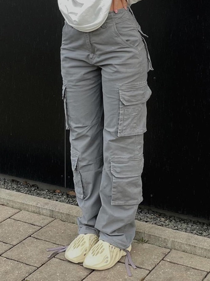 Vrouw draagt een grijze cargobroek met wijde pijpen en witte top, staand voor een zwarte muur.