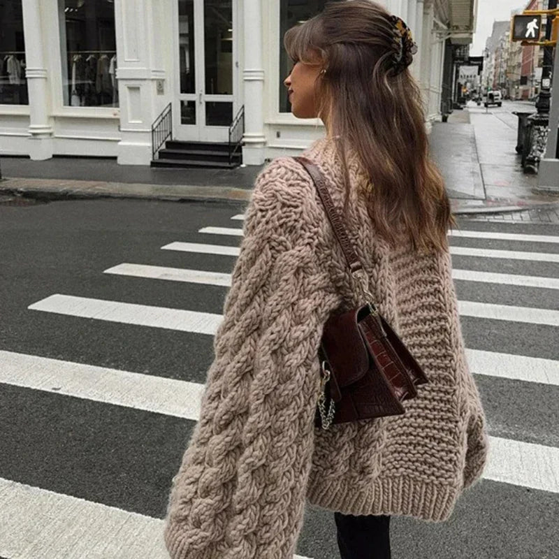 Vrouw in oversized khaki gebreide trui loopt over een zebrapad in de stad met een handtas.