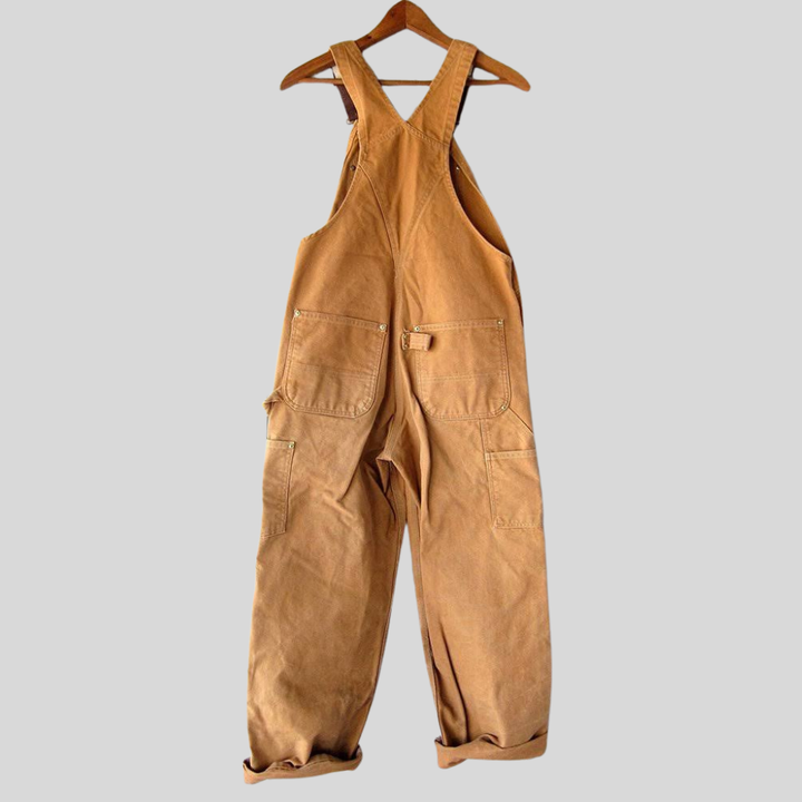 Dames retro tuinbroek in warme oranje kleur hangend aan een houten hanger, met ruime zakken en verstelbare bandjes